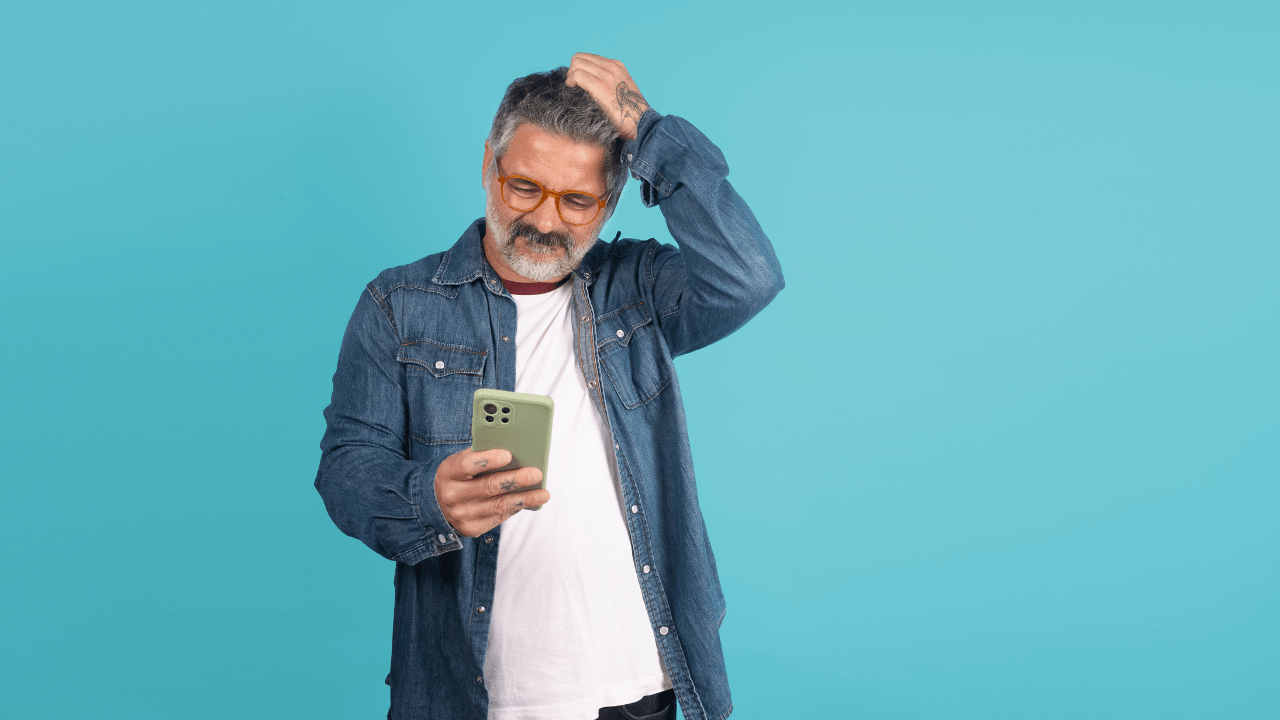 Confused man in a denim shirt looking at his phone and scratching his head against a teal background